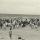 Une photographie en noir et blanc d’une foule de personnes, la plupart debout, sur une plage.